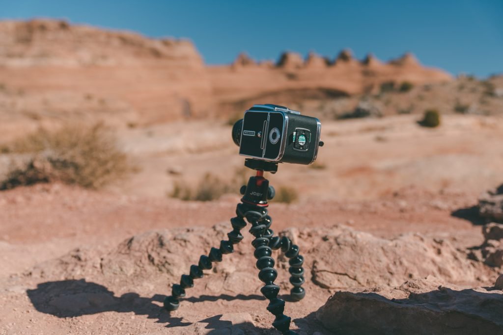 A camera mounted on a flexible Joby tripod stands on rocky terrain, with reddish desert cliffs and blue sky in the background.