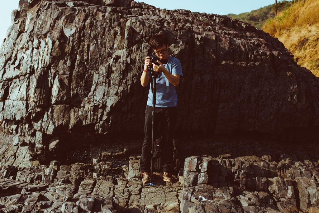 Young man with glasses adjusting a camera on a monopod, standing in front of a large, textured rock formation, with sunlit dry grasses in the background.