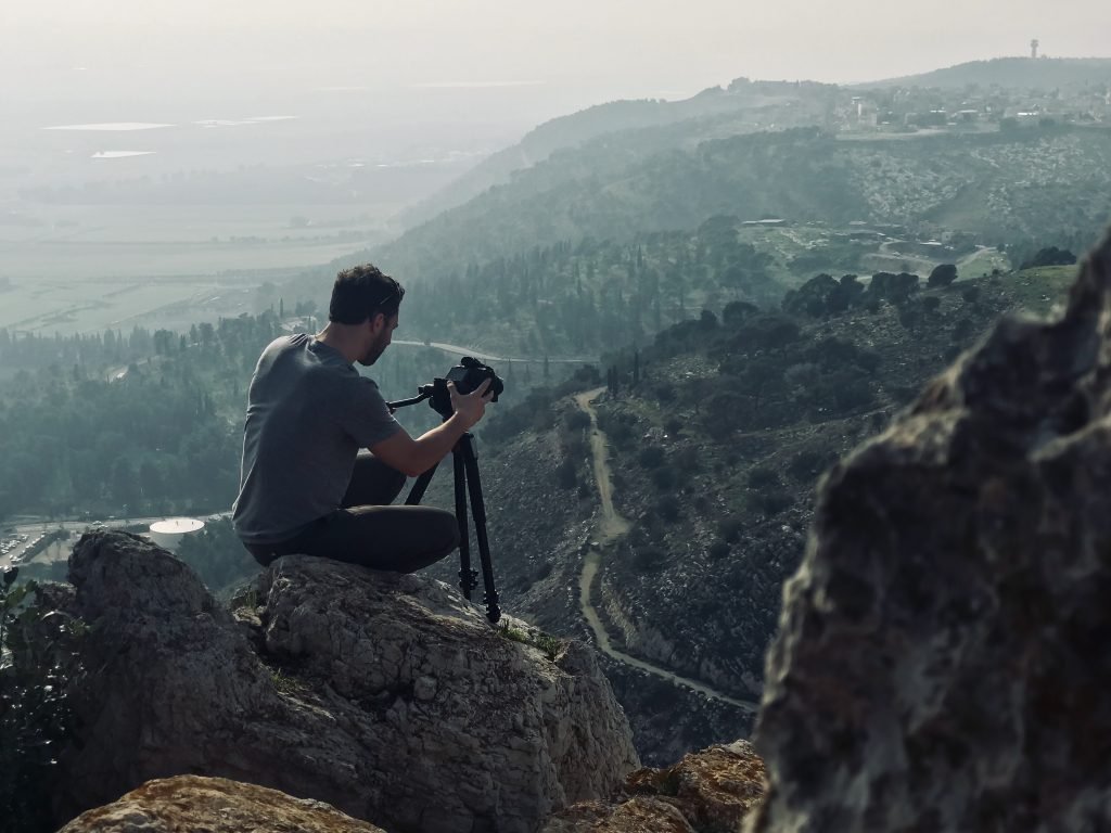 Photographer capturing the landscape from a high vantage point with a tripod, overlooking a scenic valley with winding roads and hills in the background.