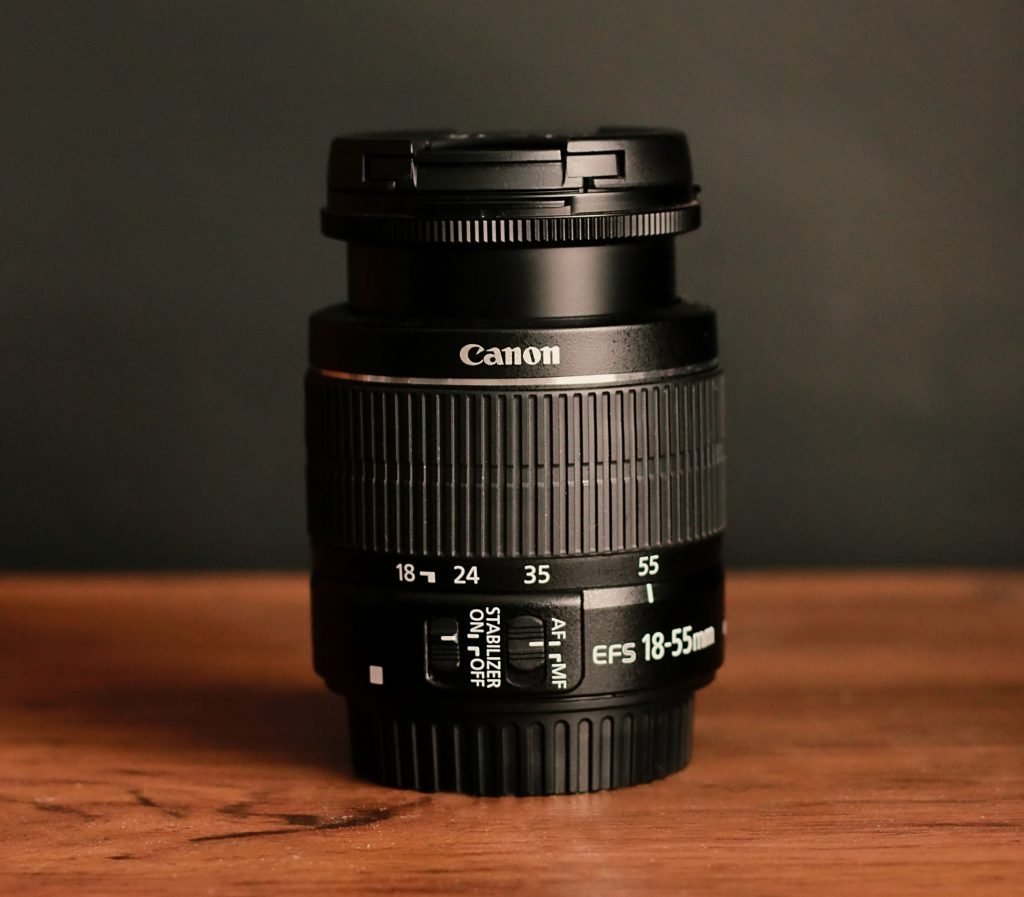 A Canon EFS 18-55mm camera lens displayed on a wooden surface with a dark background, showcasing its focus range markers and stabilization switch.