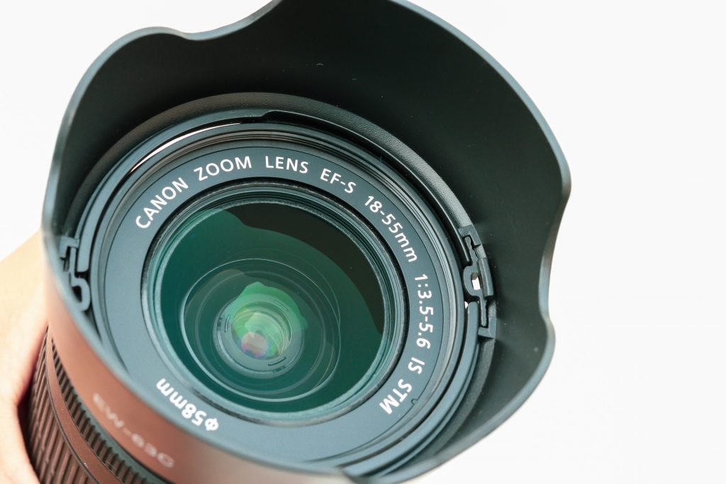 Close-up view of a Canon Zoom Lens EFS 18-55mm with its lens hood attached, showcasing the lens details and the reflective glass surface against a light background.