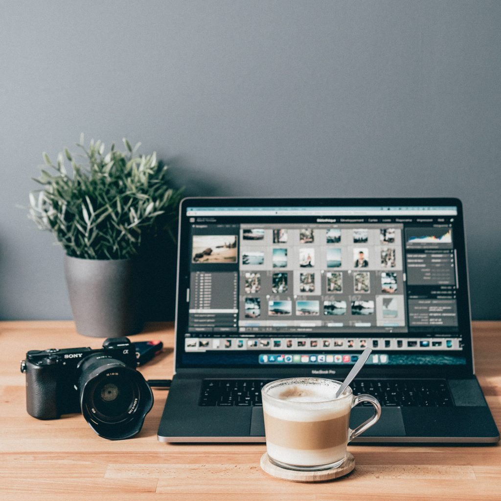 Modern photographer's workspace with a Sony camera, a laptop displaying photo editing software, a fresh cup of coffee, and a potted plant on a wooden desk.