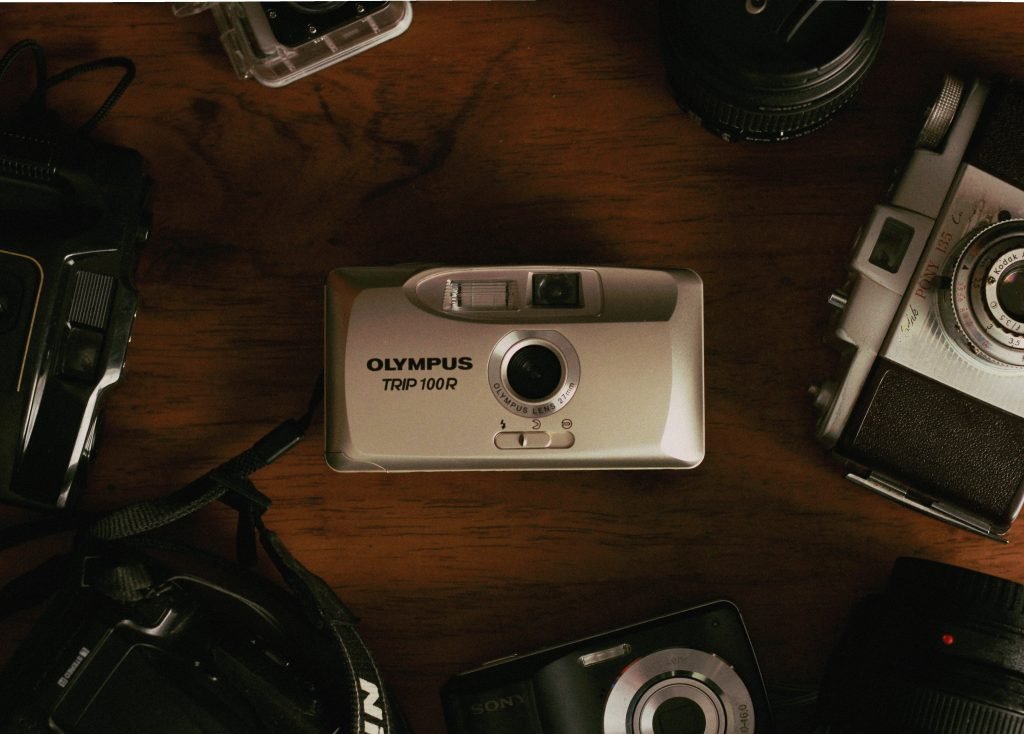 An assortment of cameras and photography equipment scattered on a wooden surface. Prominently in the center is an Olympus Trip 100R film camera. Surrounding it are parts of other cameras and accessories, including a camera lens to the right and parts of other camera bodies in the surrounding area, indicative of a photographer's workspace or collection.