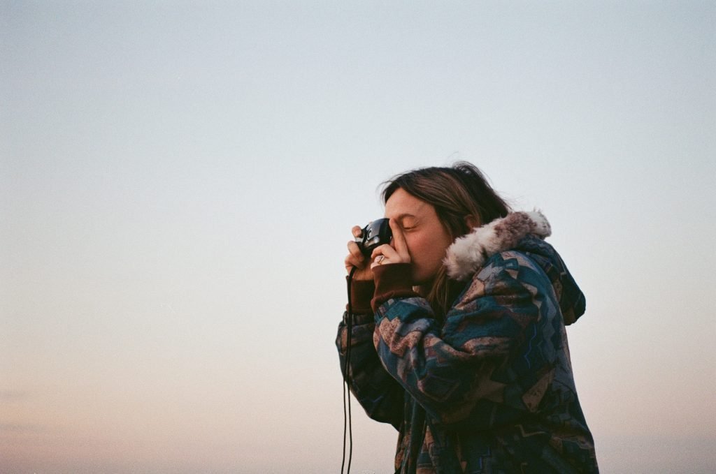 A girl with shoulder-length hair is seen taking a photo with a vintage camera, their eye pressed to the viewfinder. They are wearing a patterned jacket with a fur-lined hood. The background is a soft, pastel-colored sky suggesting either dawn or dusk. The focus on the individual against the expansive sky conveys a sense of solitude and the quiet focus associated with photography.