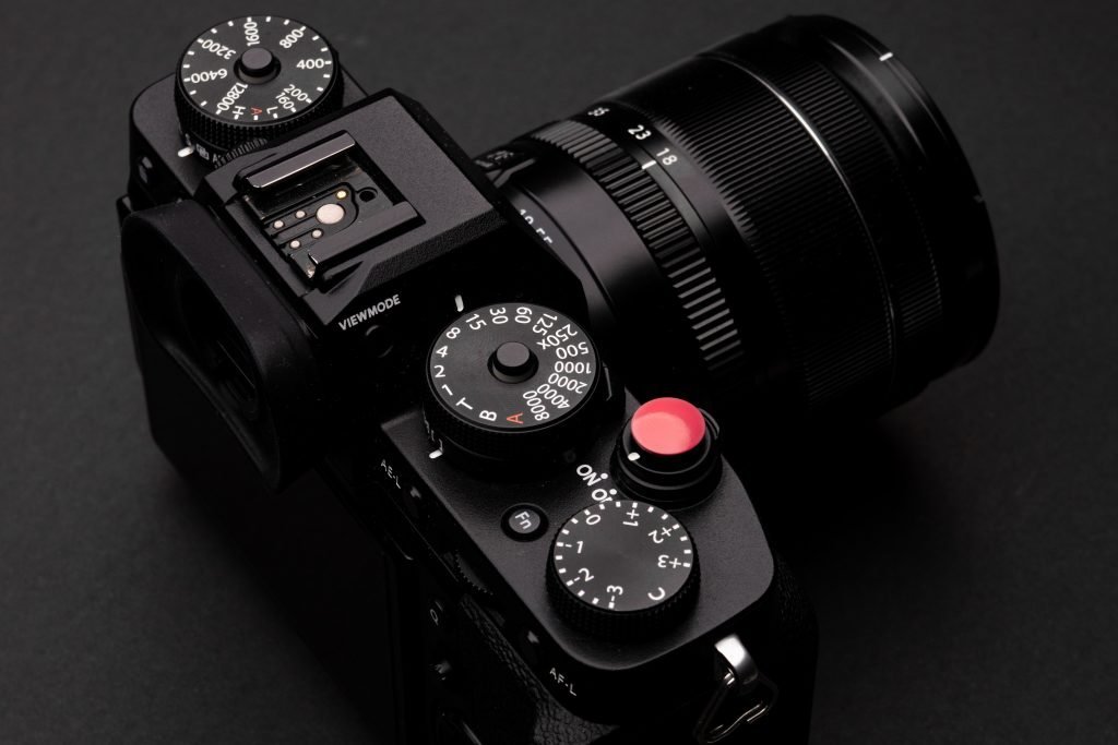 This is a close-up image of a modern digital camera with a black body and a lens attached. The top of the camera shows various dials and buttons, including one with a red dot, typically used for starting and stopping video recording or taking photographs. There's a shutter speed dial, an exposure compensation dial, and a mode selection dial visible, indicating the camera's manual functionality. The camera appears to be a high-end, professional model with intricate controls, designed for precision photography. The black background contrasts with the camera, highlighting its details and design.