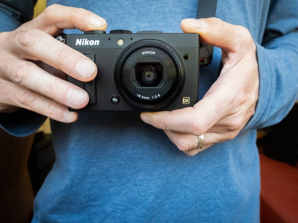 Close-up of a person's hands holding a Nikon compact camera with a fixed lens, ready to take a photo. The camera is positioned centrally, with the lens facing directly towards the viewer.